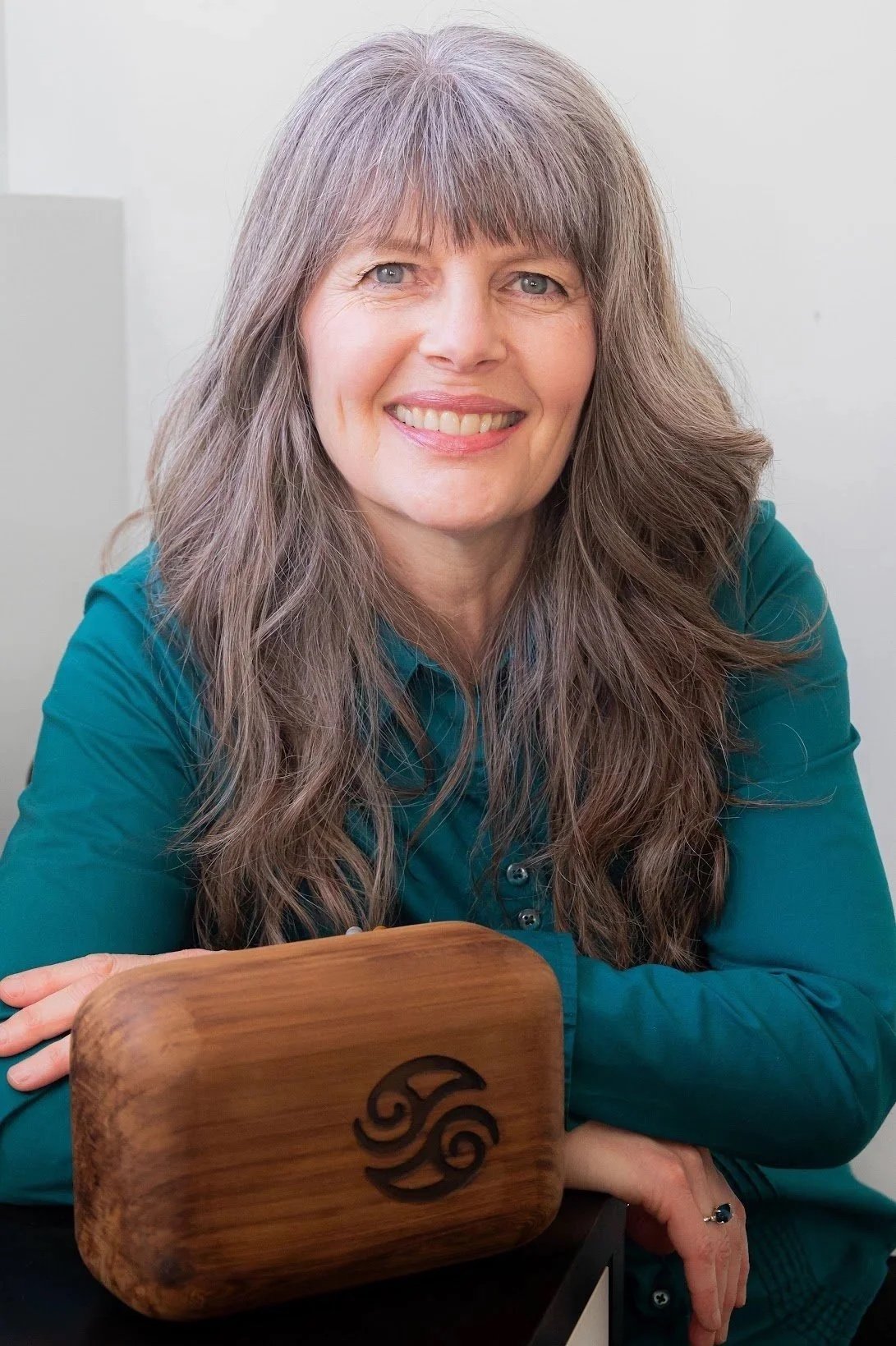 A woman with long gray hair and blue eyes sitting at a table, smiling, with her arms resting on a wooden block with a carved logo.
