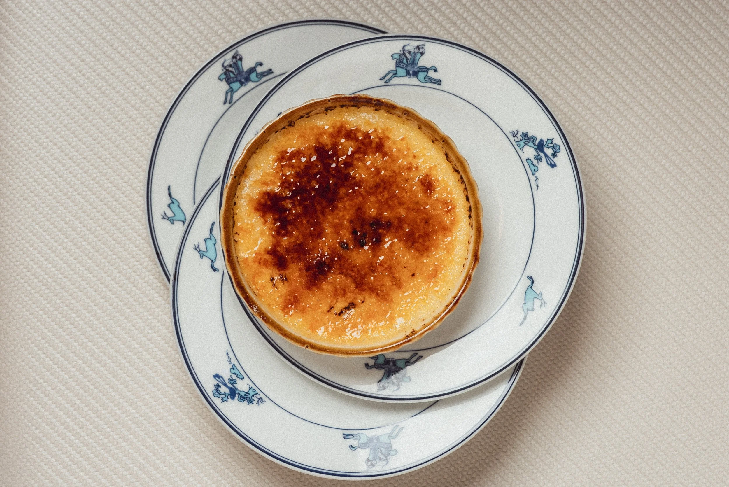 A dessert, possibly crème brûlée, served in a small ceramic dish with a caramelized sugar crust on top, placed on a decorative blue and white plate with animal designs.