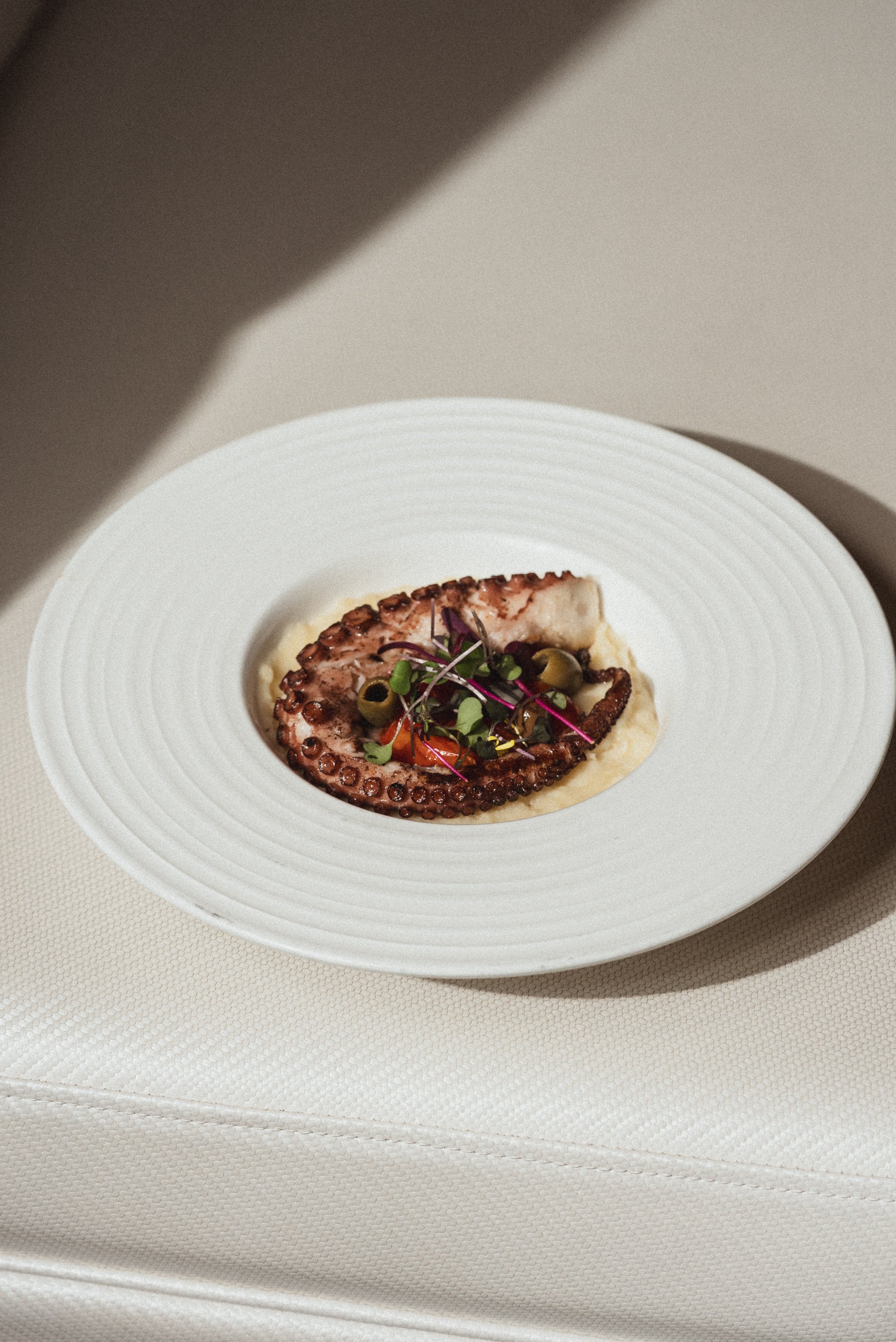 A plate of cooked octopus tentacle garnished with microgreens and olives, served with mashed potatoes.
