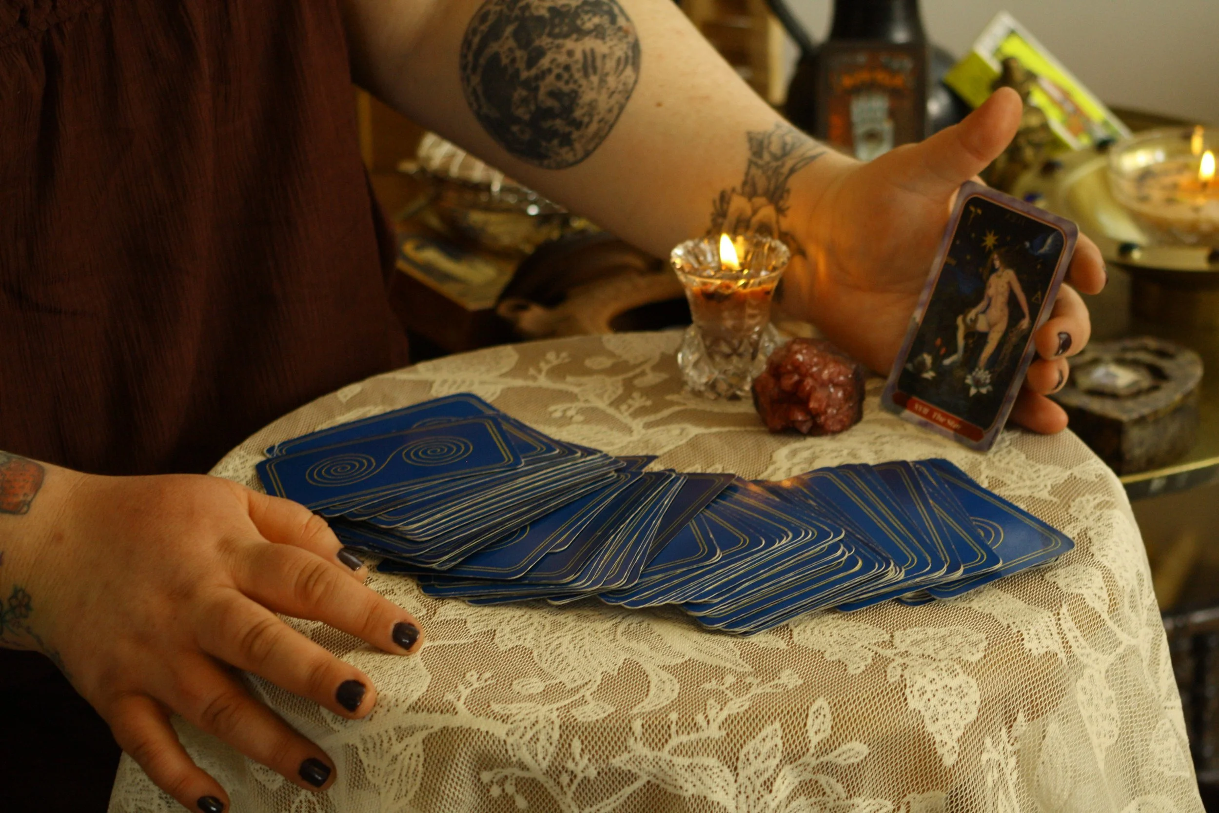 A person with tattoos on their arm holding a tarot card in one hand and a deck of tarot cards in the other. There are lit candles, crystals, and tarot-themed items on a table covered with a lace tablecloth.