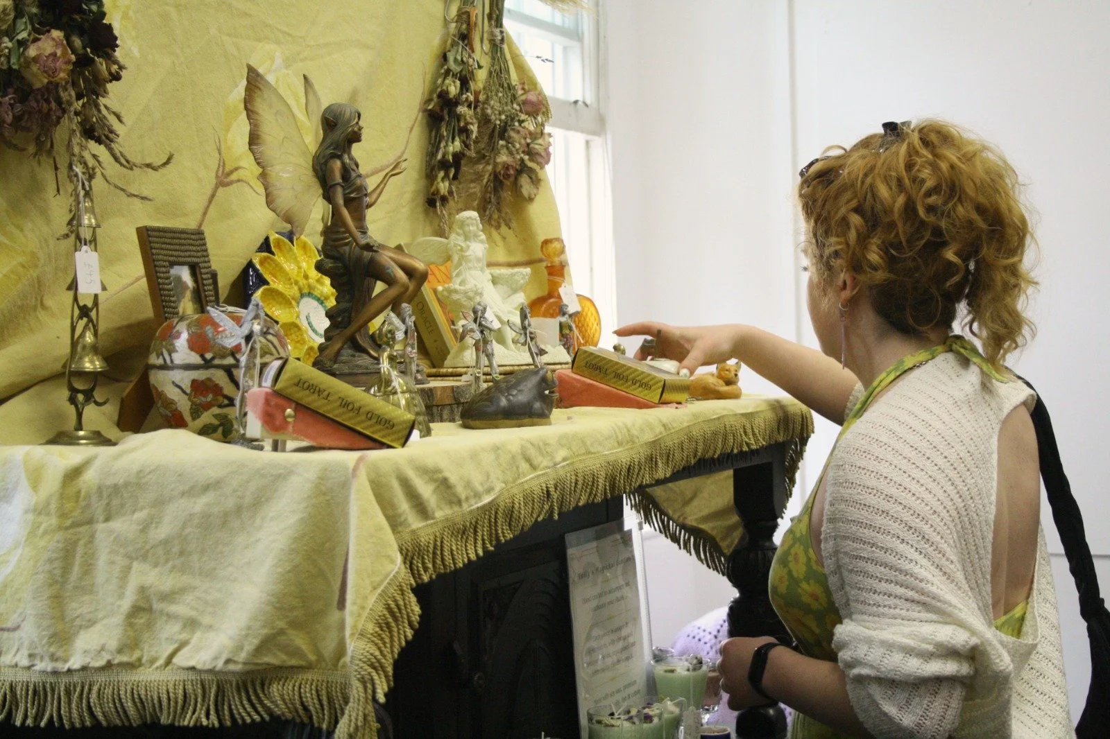 A woman with curly red hair and earrings, wearing a sleeveless top and a crocheted cover-up, looks at a display of decorative figurines, books, and dried flowers on a yellow cloth-covered table in a bright room.