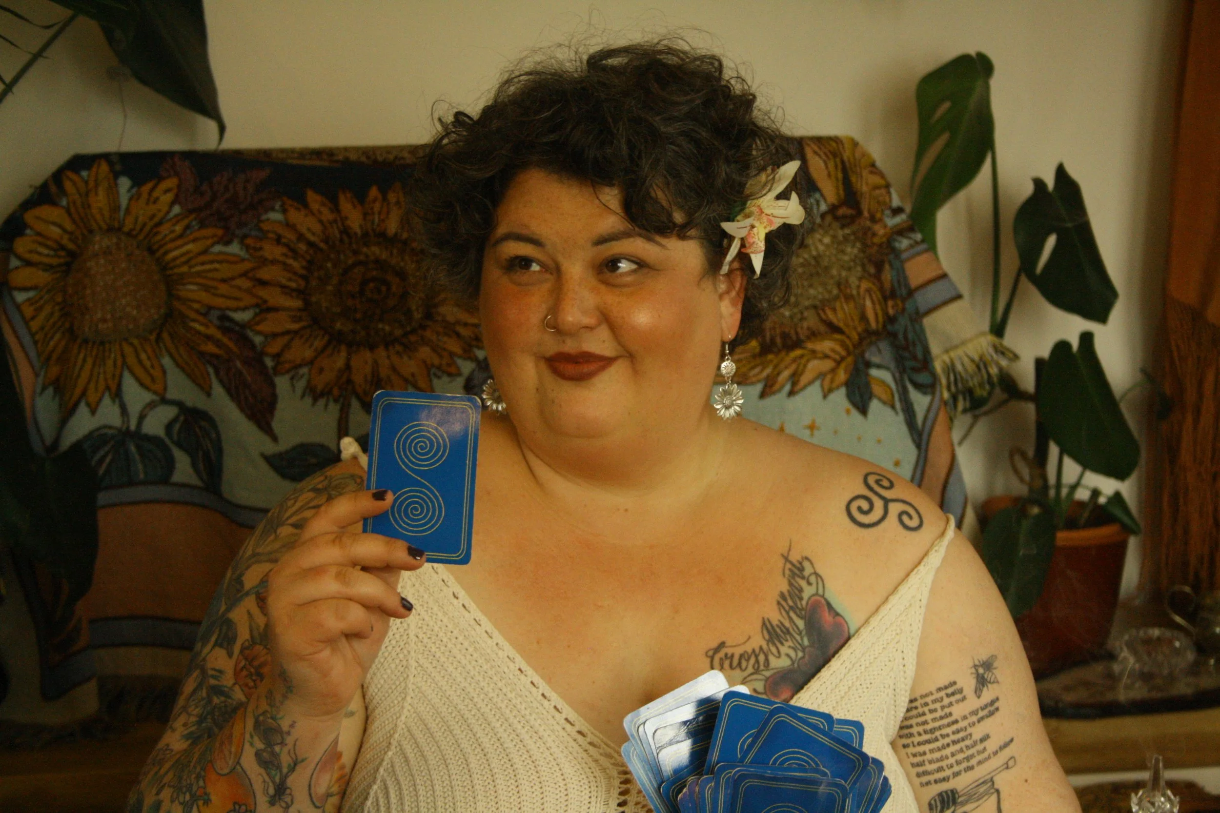 A woman with short curly dark hair, tattoos, earrings, and a flower in her hair, holding tarot cards in a room with plants and a sunflower-patterned blanket.