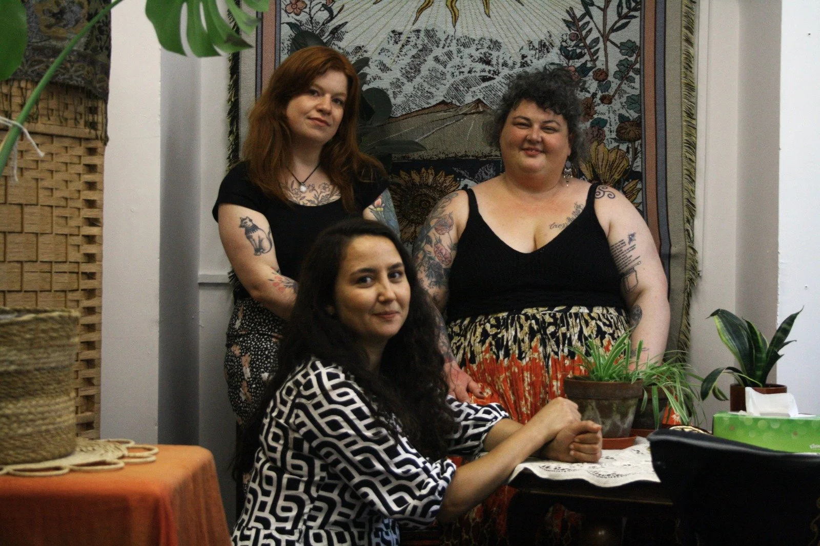 Three women with tattoos, sitting in the tarot reading space at Altar & Arcana. They are in a room decorated with plants and a tapestry. They are smiling and looking at the camera.