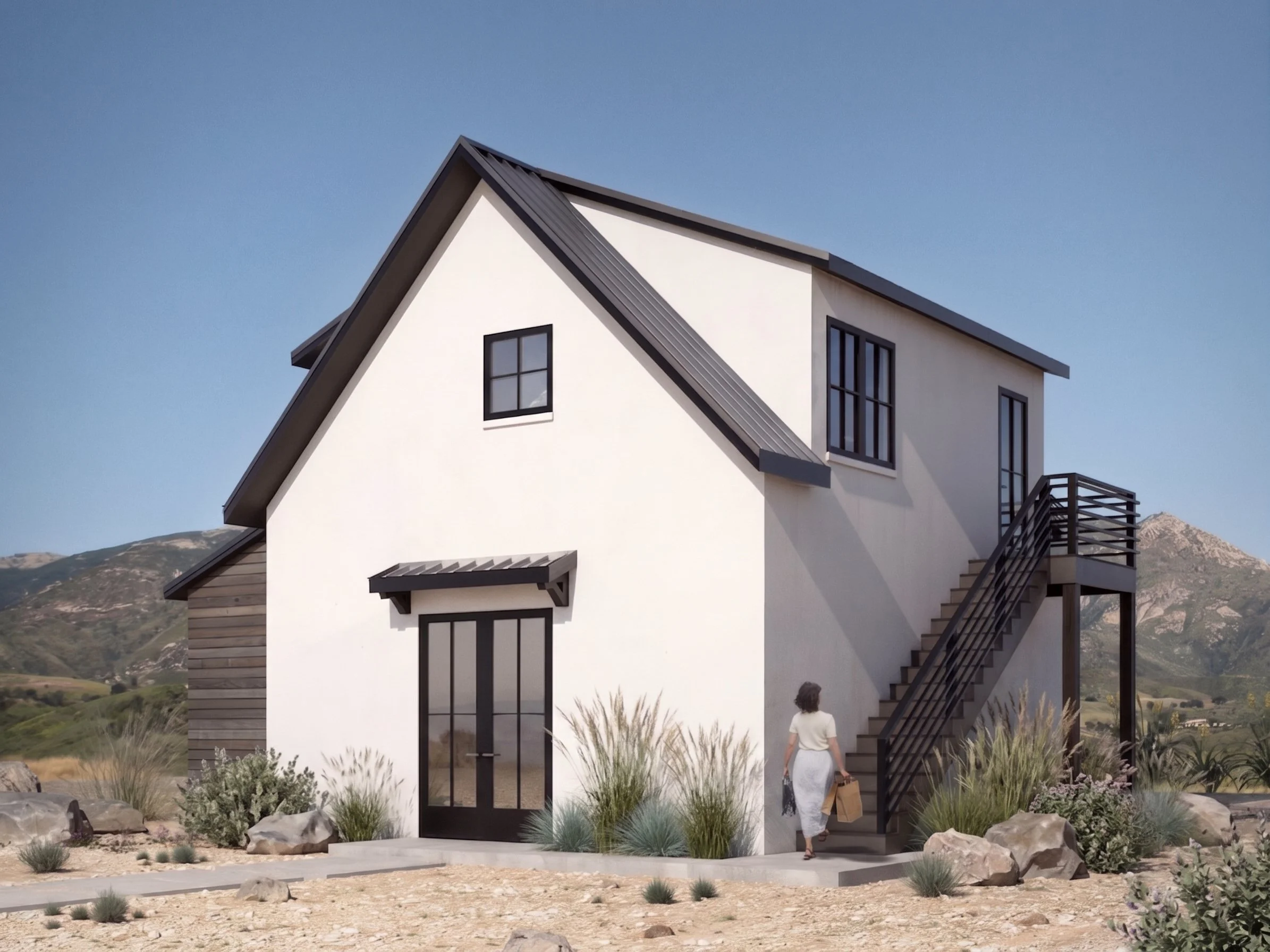 Modern white house with black trim, metal roof, and exterior stairs, set in a landscape with rocks, grasses, and mountains in the background.