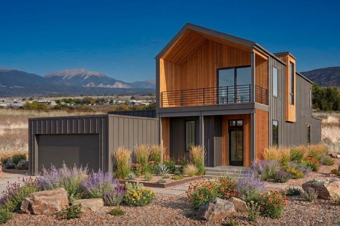 Modern two-story house with wooden and metal exterior, large windows, and a balcony, surrounded by desert landscaping with rocks, shrubs, and flowers, mountains in the background.