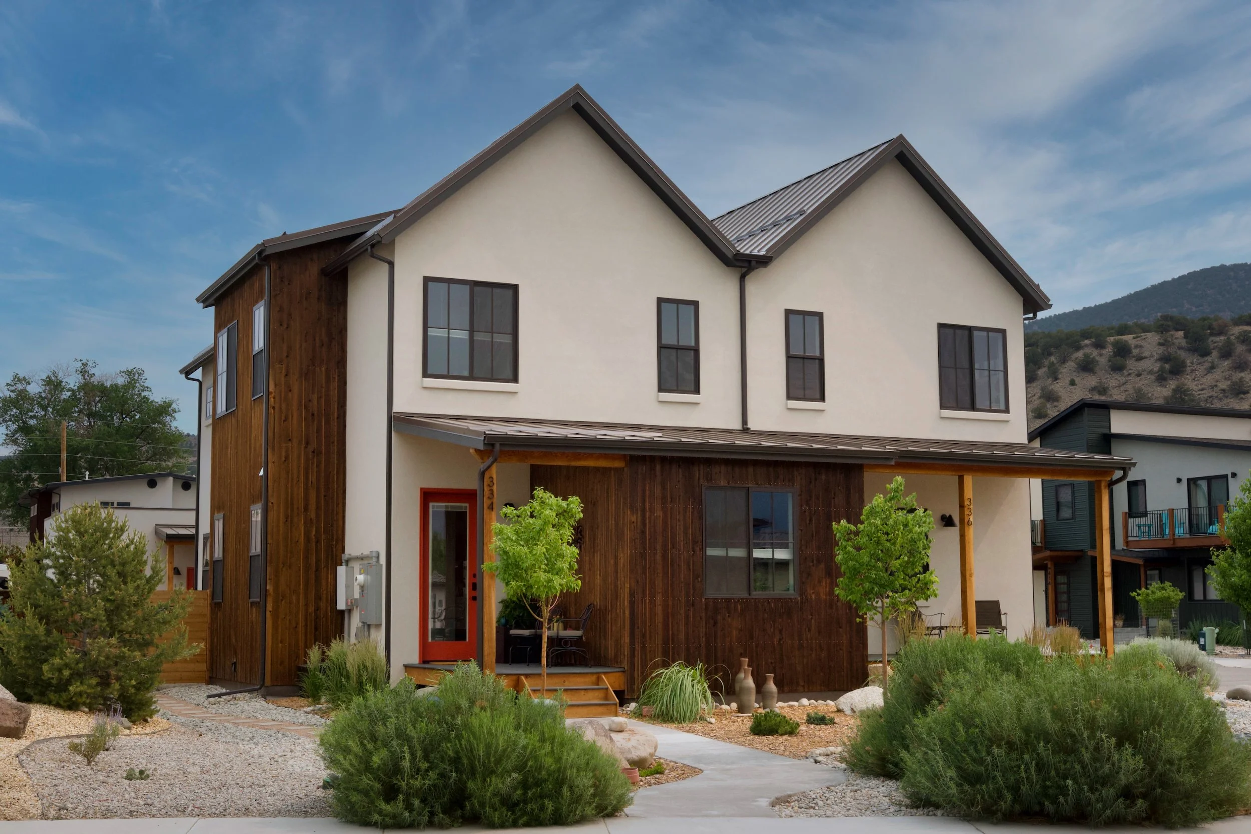 Modern two-story house with a white facade, wooden accents, and large black-framed windows, surrounded by a landscaped yard with trees and shrubs, and mountains in the background under a cloudy sky.