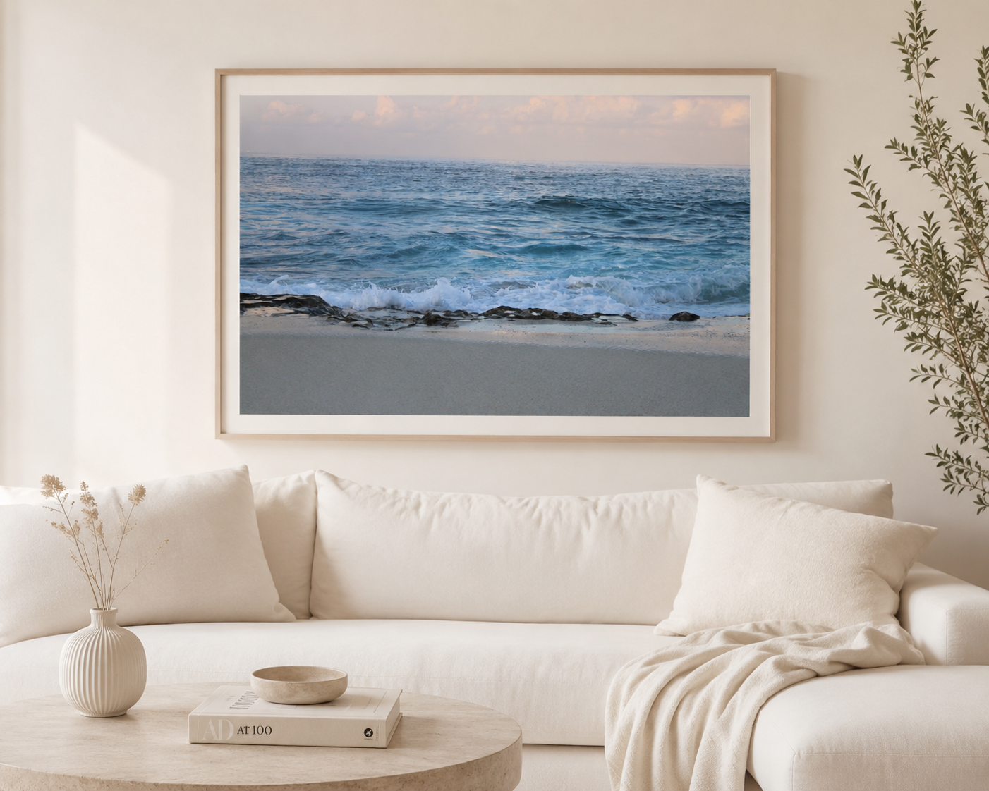 A framed photograph of the ocean with waves crashing onto a sandy beach hung on a light-colored wall above a white sofa. There is a potted plant on the right and a small round table with books, a bowl, and a vase on the left.