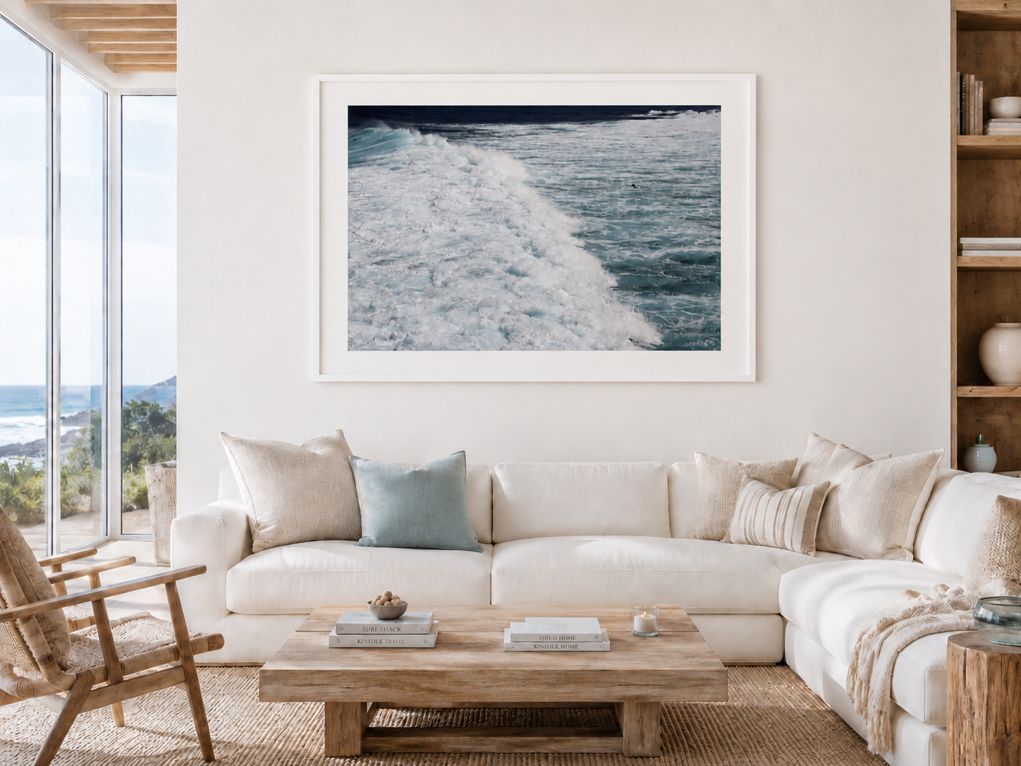 Living room with white sofa, pillows, wooden coffee table, wall art of ocean waves, and glass doors showing a coastal landscape.