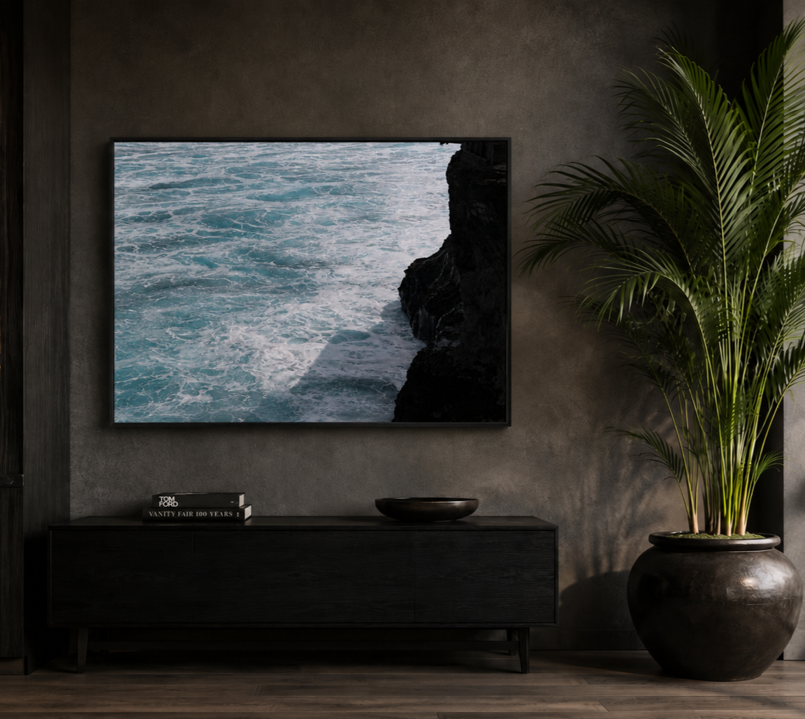 Interior of a modern living room with a large framed ocean and rock photograph on a gray wall, a black console table with a black bowl and two books, and a tall potted plant with large green leaves.