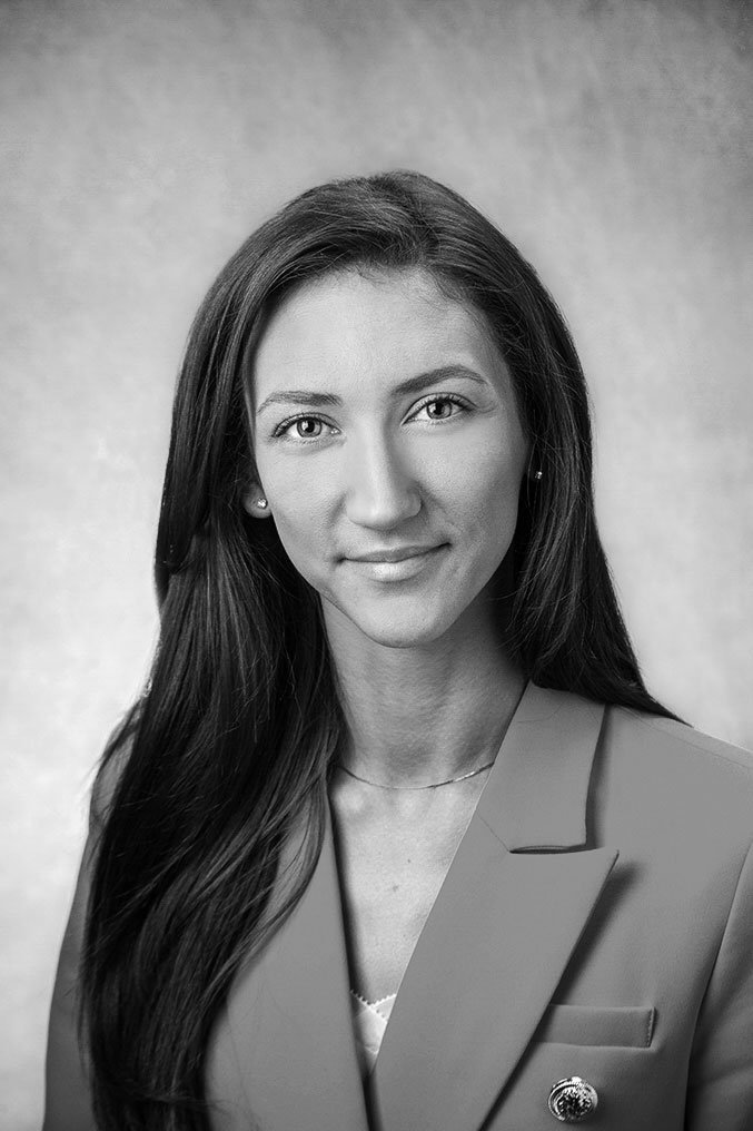 Black and white portrait of a woman with long dark hair, wearing a blazer, a light top, and a necklace, smiling slightly.