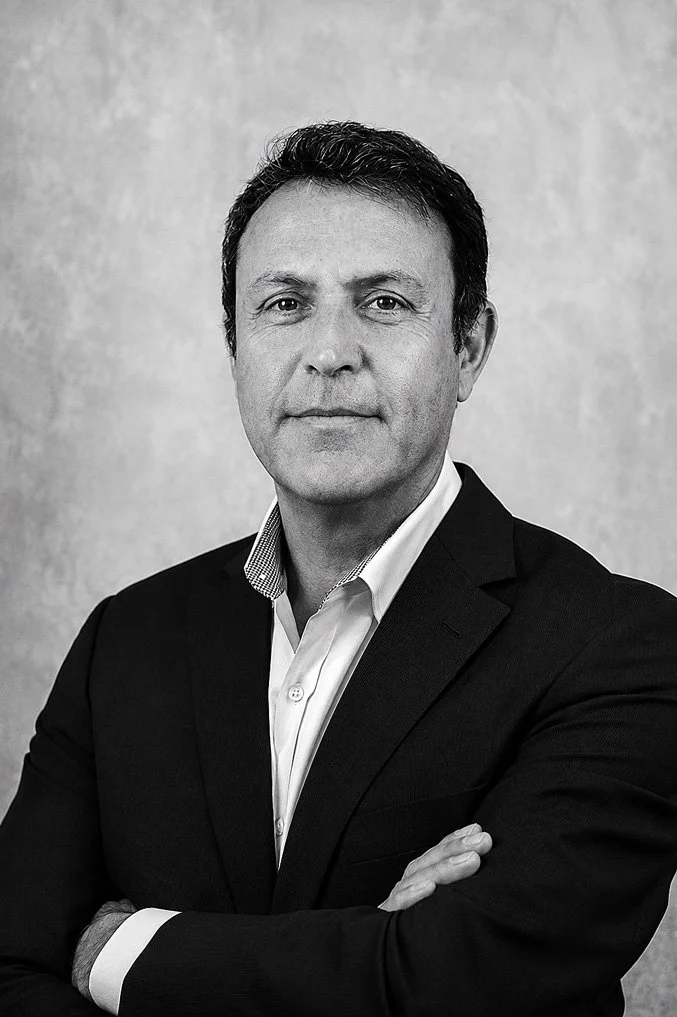 Black and white professional portrait of a middle-aged man with dark hair, wearing a suit and collared shirt, standing with arms crossed in front of a plain background.