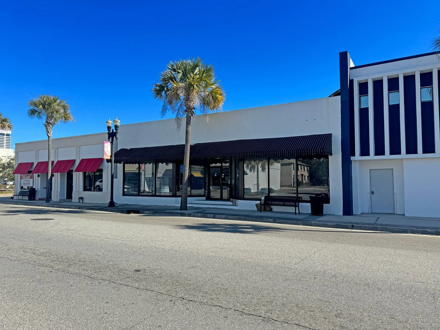 A small storefront with large glass windows, black awning, and red awnings over the side windows, flanked by palm trees and a blue sky.