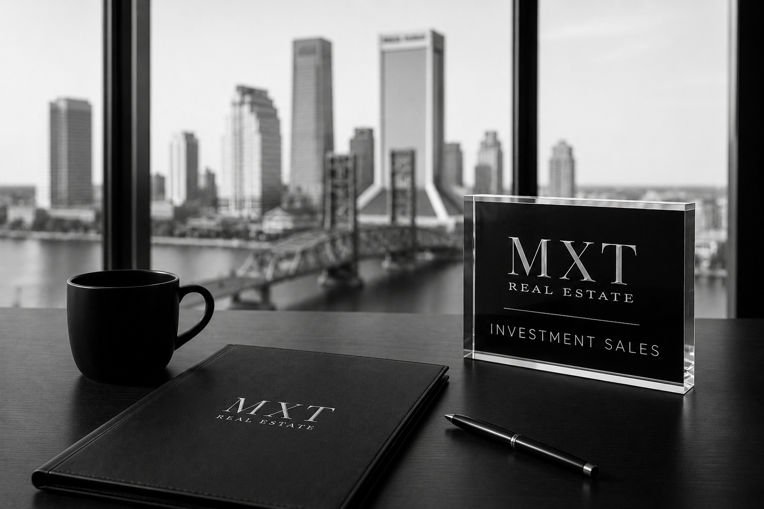 A black desk with a mug, a closed planner, a pen, and a glass sign that reads 'MXT Real Estate Investment Sales,' with a city skyline view of skyscrapers and a bridge through large windows in the background.