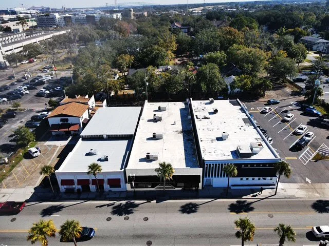 Aerial view of a commercial building with a parking lot, surrounded by trees, other parking areas, and cityscape in the background.