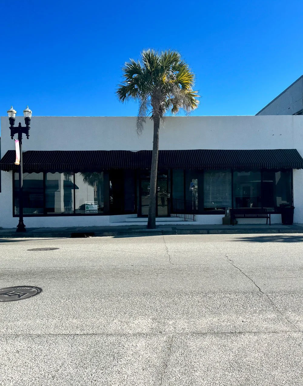 A white building with large front windows and a black striped awning, a palm tree in front, a bench to the right, a lamp post to the left, and a clear blue sky.