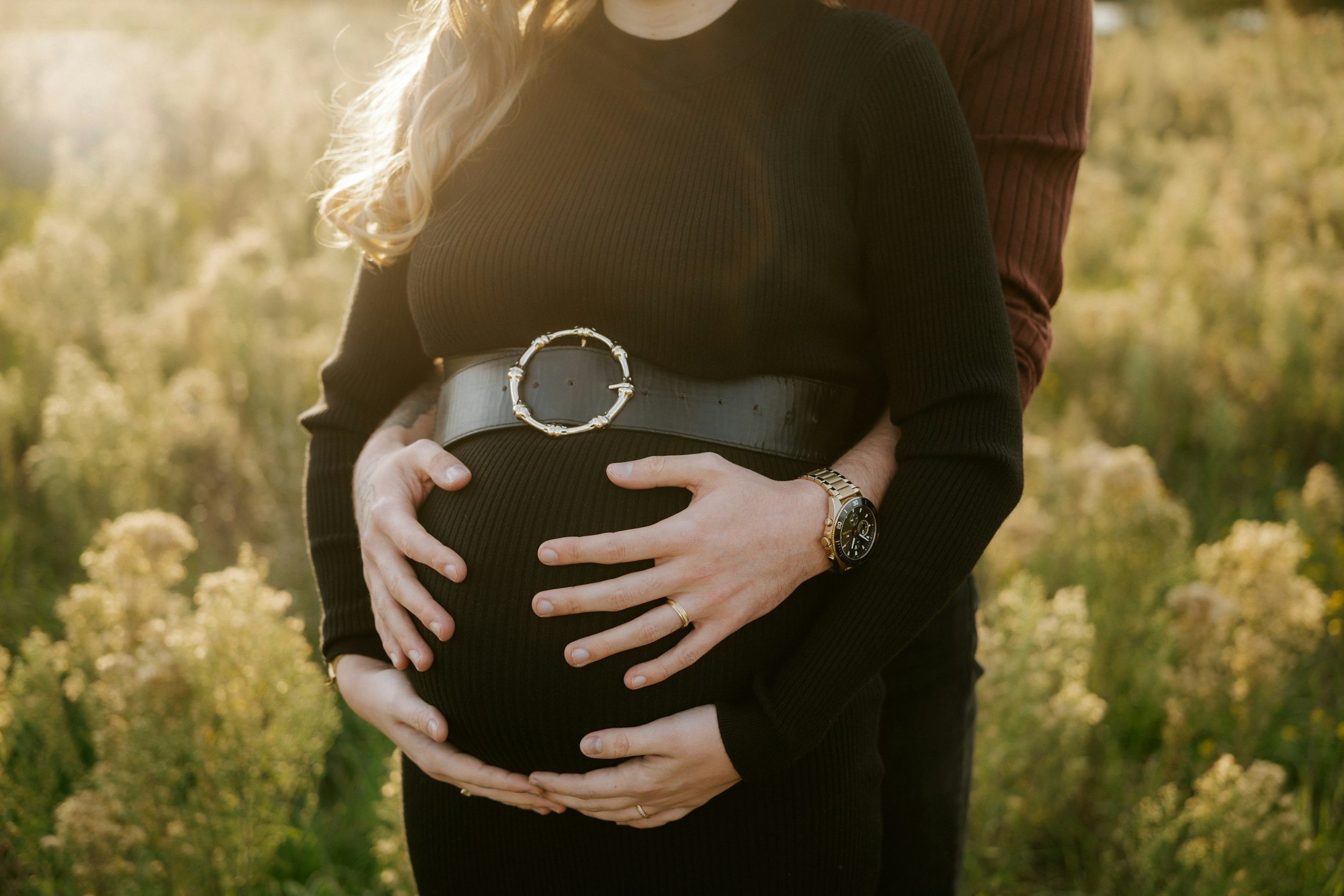 A pregnant woman in a black long-sleeve top with a wide belt, standing outdoors with sunlight, with her hands on her belly, and a partner behind her with one arm around her shoulder. The background features trees and yellow flowers.