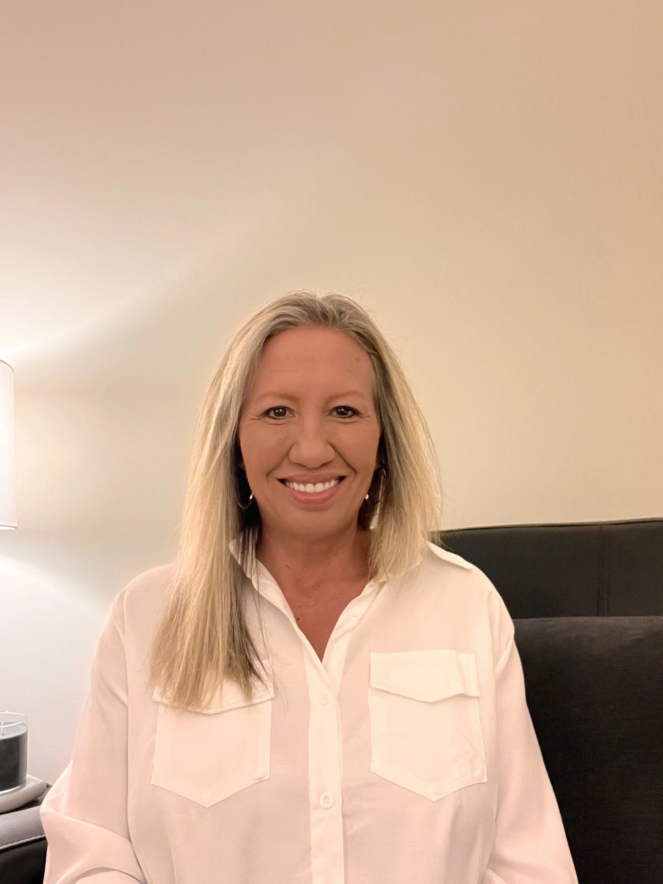 A woman with long blonde hair smiling and wearing a light-colored blouse, sitting in a room with plain walls and a black piece of furniture behind her.