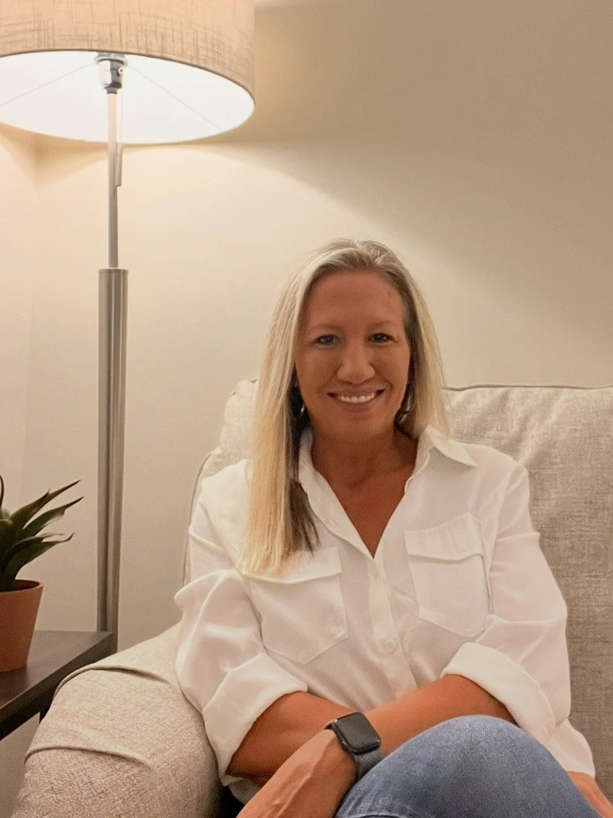 A grief therapist in Overland Park, Kansas  with long blonde hair smiling, sitting on a beige armchair in a room with a tall floor lamp and a potted plant beside her.