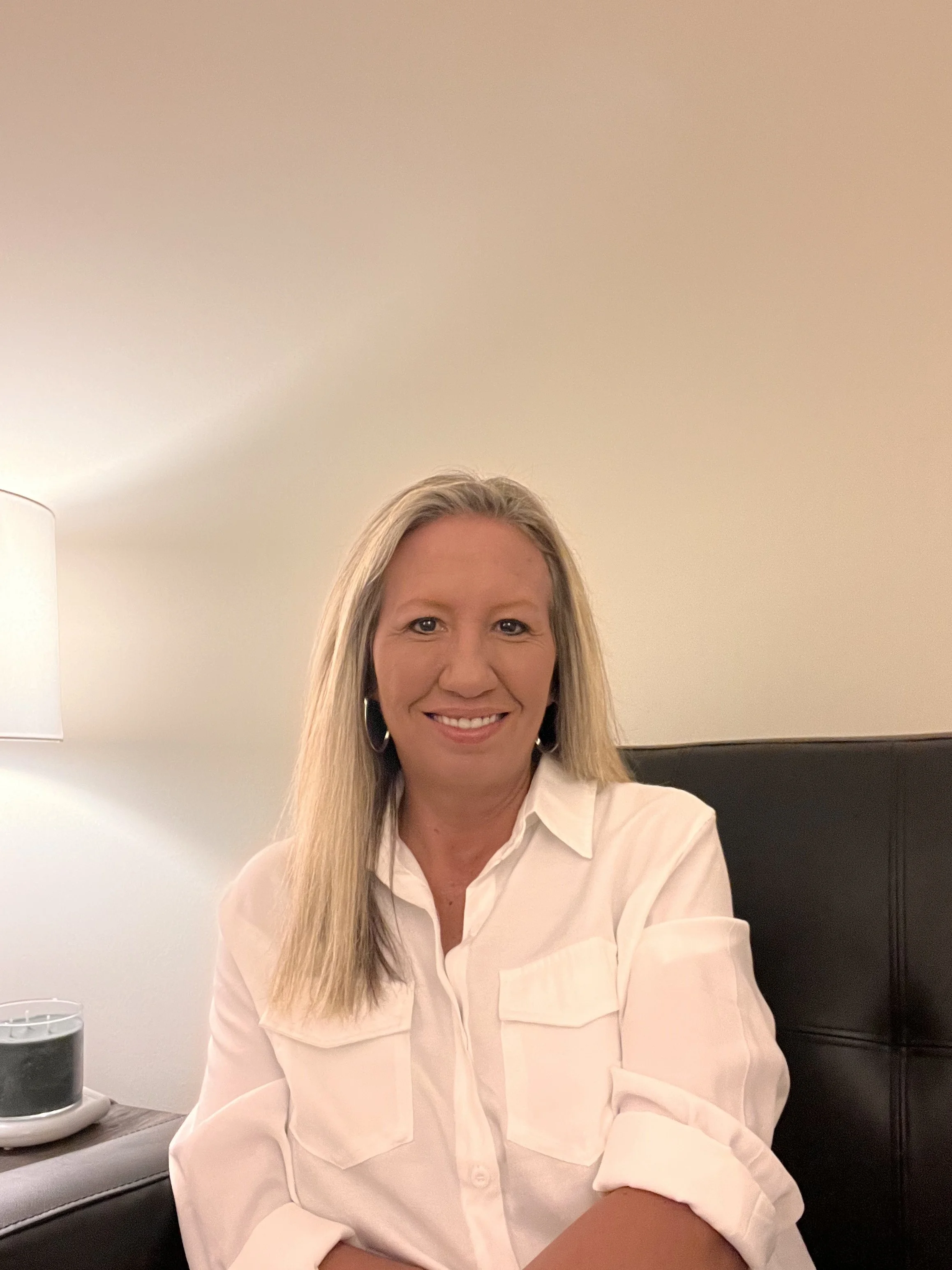 A smiling woman with long blonde hair, wearing a white blouse, sitting on a black headboard in a room with beige walls, with a lamp and a small black object on the nightstand beside her.