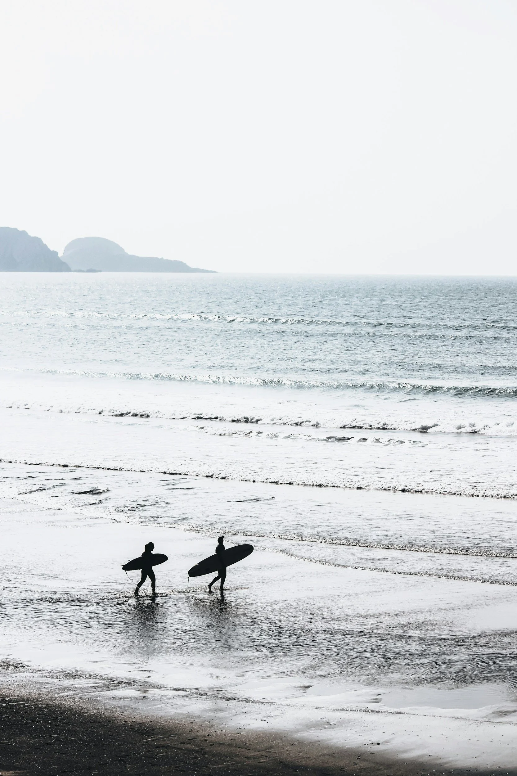 Silhouettes of two surfers walking along a beach with surfboards, with the ocean and distant cliffs in the background during sunset or sunrise.
