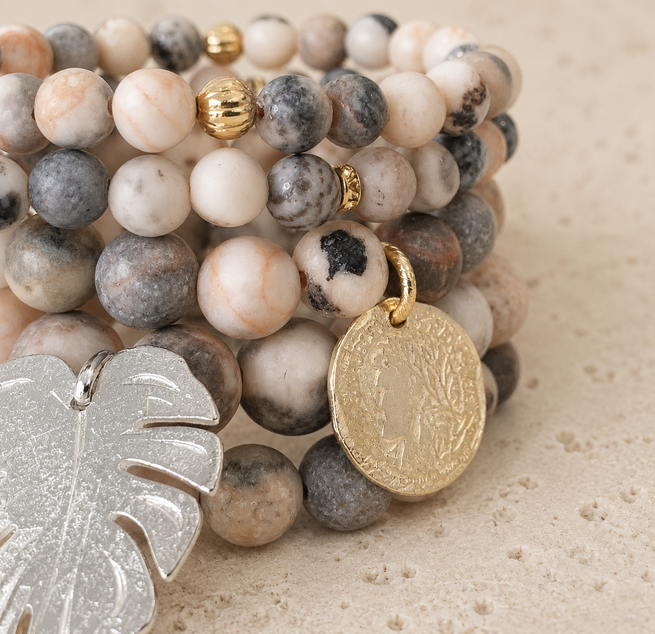 Close-up of beaded jewelry with round stone beads in black, white, and gray, accented with gold and silver charms, including a gold coin and a silver leaf-shaped pendant, on a beige textured surface.
