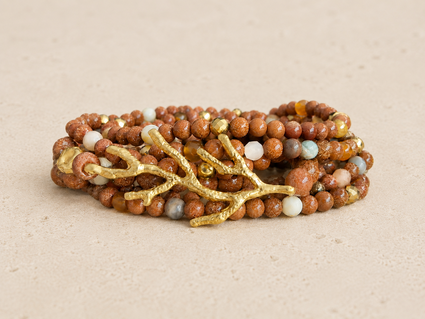 A multi-layered bracelet made of brown, white, and colorful beads with gold branch and leaf accents, resting on a beige surface.