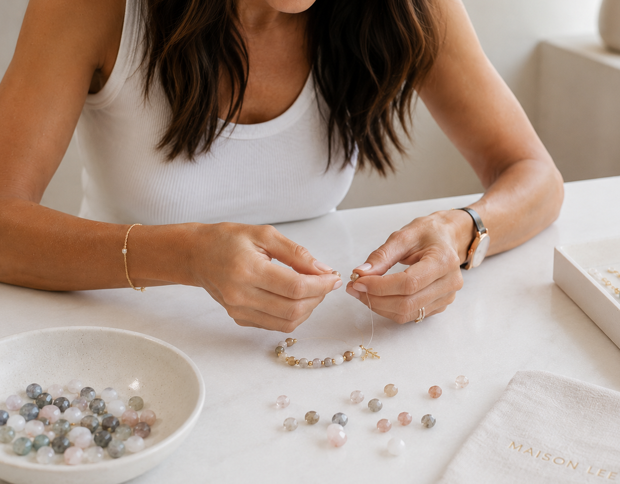 A woman assembling a beaded bracelet at a white table, with a bowl of multicolored beads, loose beads, and jewelry-making supplies nearby.