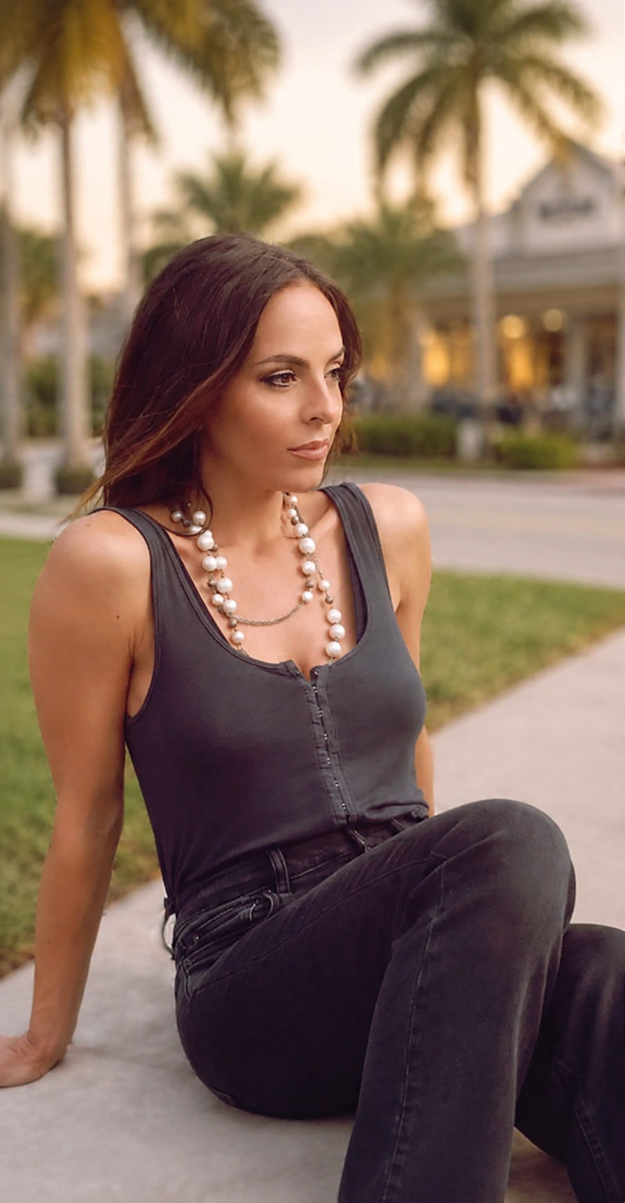 A woman with brown hair and a necklace, wearing a black tank top and black jeans, sitting on a sidewalk with palm trees in the background during sunset.