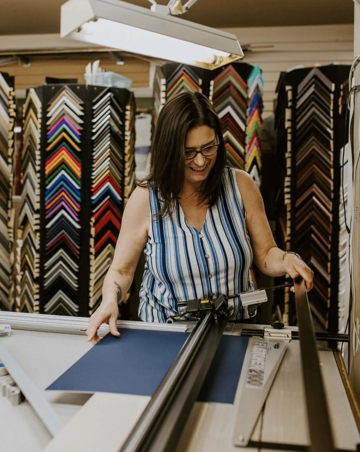 A woman working in a frame shop with colorful picture frames on racks behind her, smiling as she operates a matt cutter.