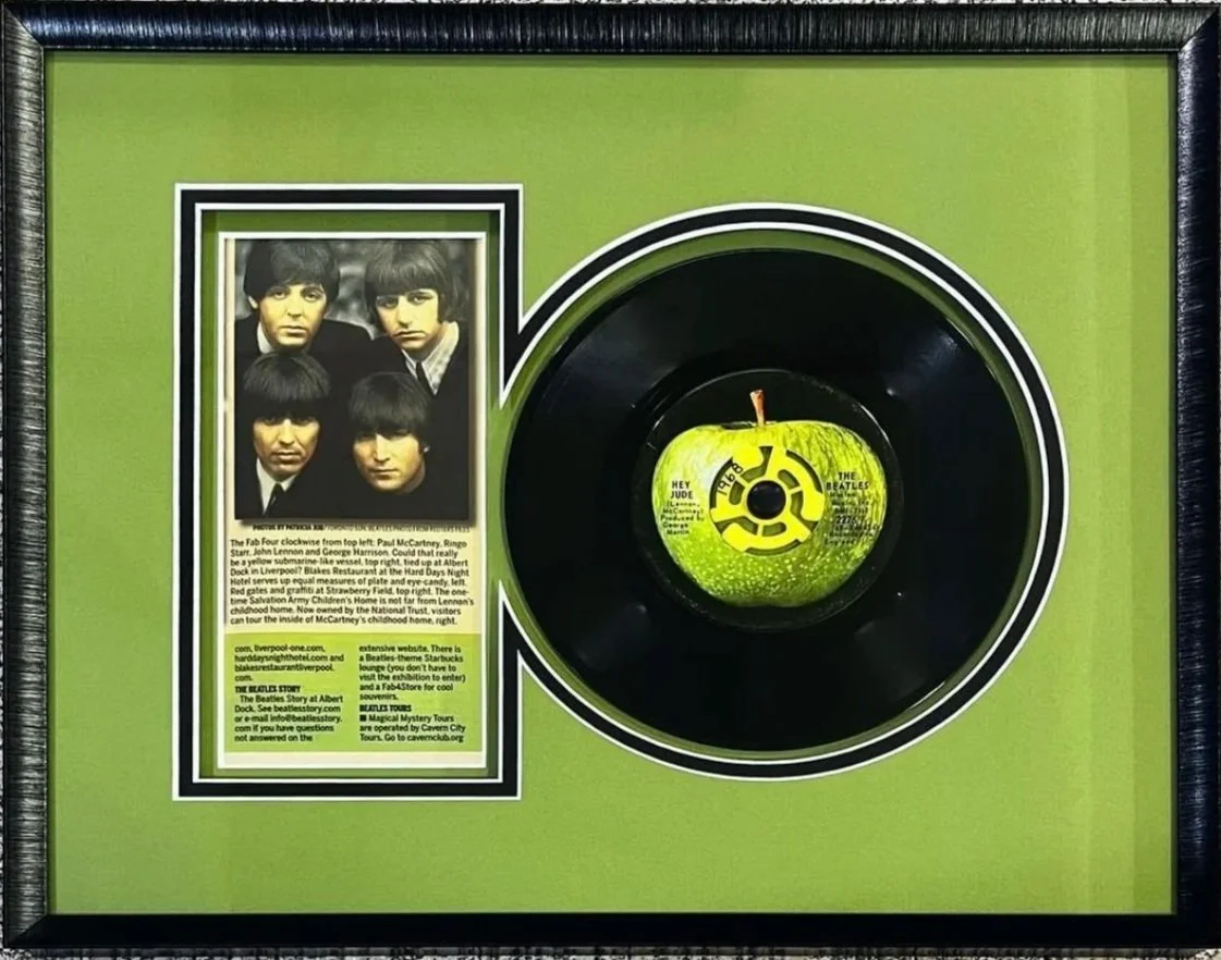 A framed display featuring a black 45 rpm record with a green apple on top, decorated with The Beatles' logo. The left side contains a black-and-white photograph of The Beatles band members, along with descriptive text.