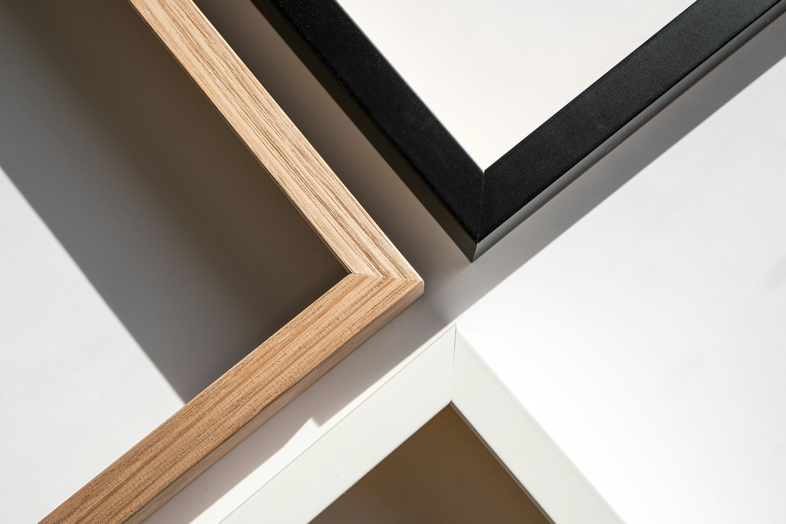 Close-up of a corner of a black picture frame, a wooden frame, and a white surface with shadows cast on it.