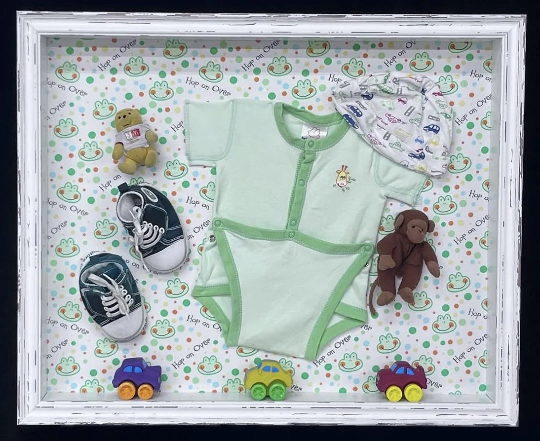 Baby clothing and toys in floater frame glass display case with patterned background.