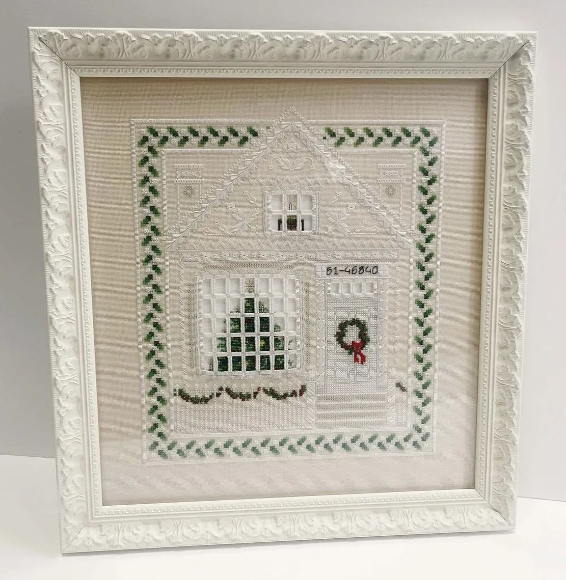 Cross-stitched picture of a house with Christmas decorations, framed in an ornate white frame.