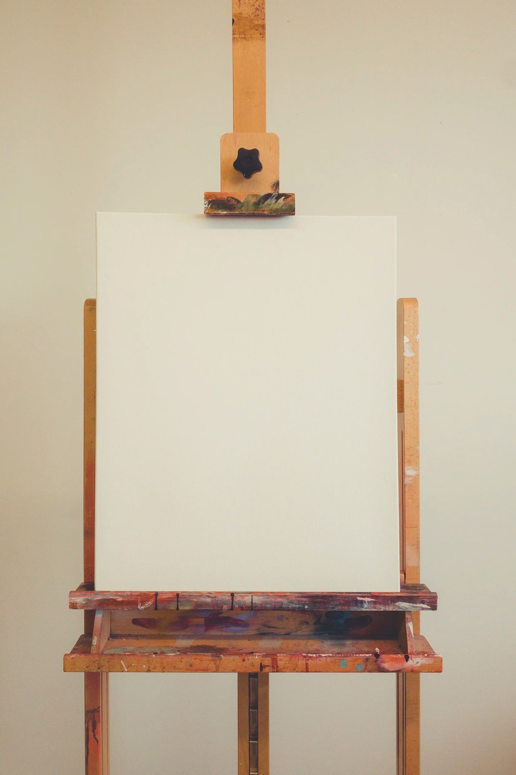 An empty white canvas on an easel in a room with beige walls, with paint splatters on the lower part of the canvas and the wooden parts of the easel.