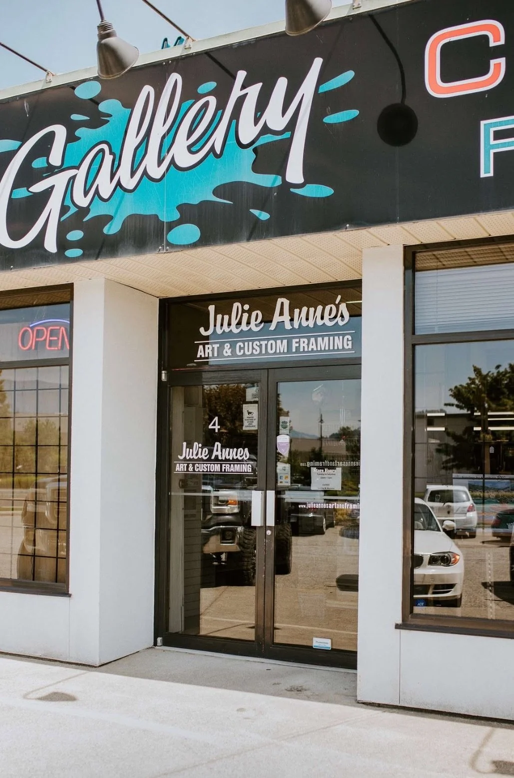 Storefront with signs reading 'Gallery,' 'Julie Annes Art & Custom Framing,' and an 'Open' sign in the window, parked cars reflected in glass doors and windows.