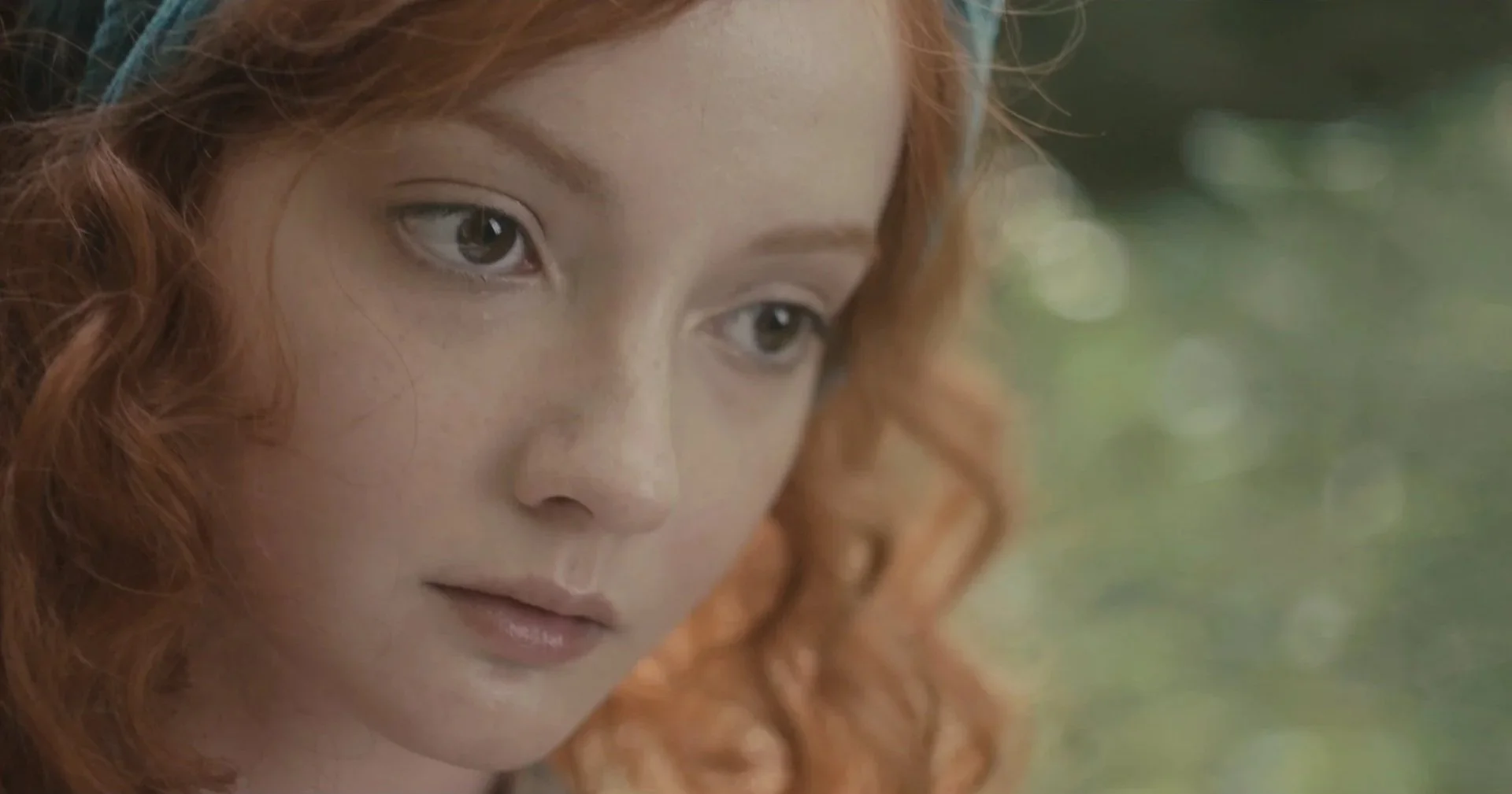 Close-up of a woman with curly red hair and green eyes, looking to the side, with a blurred outdoor background.