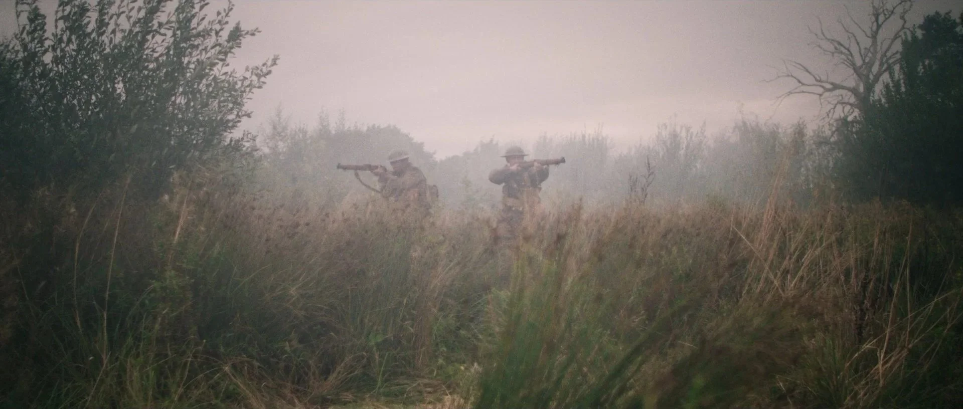 Two soldiers in military uniform walking through tall grass and bushes, aiming rifles forward, with misty weather and leafless trees in the background.