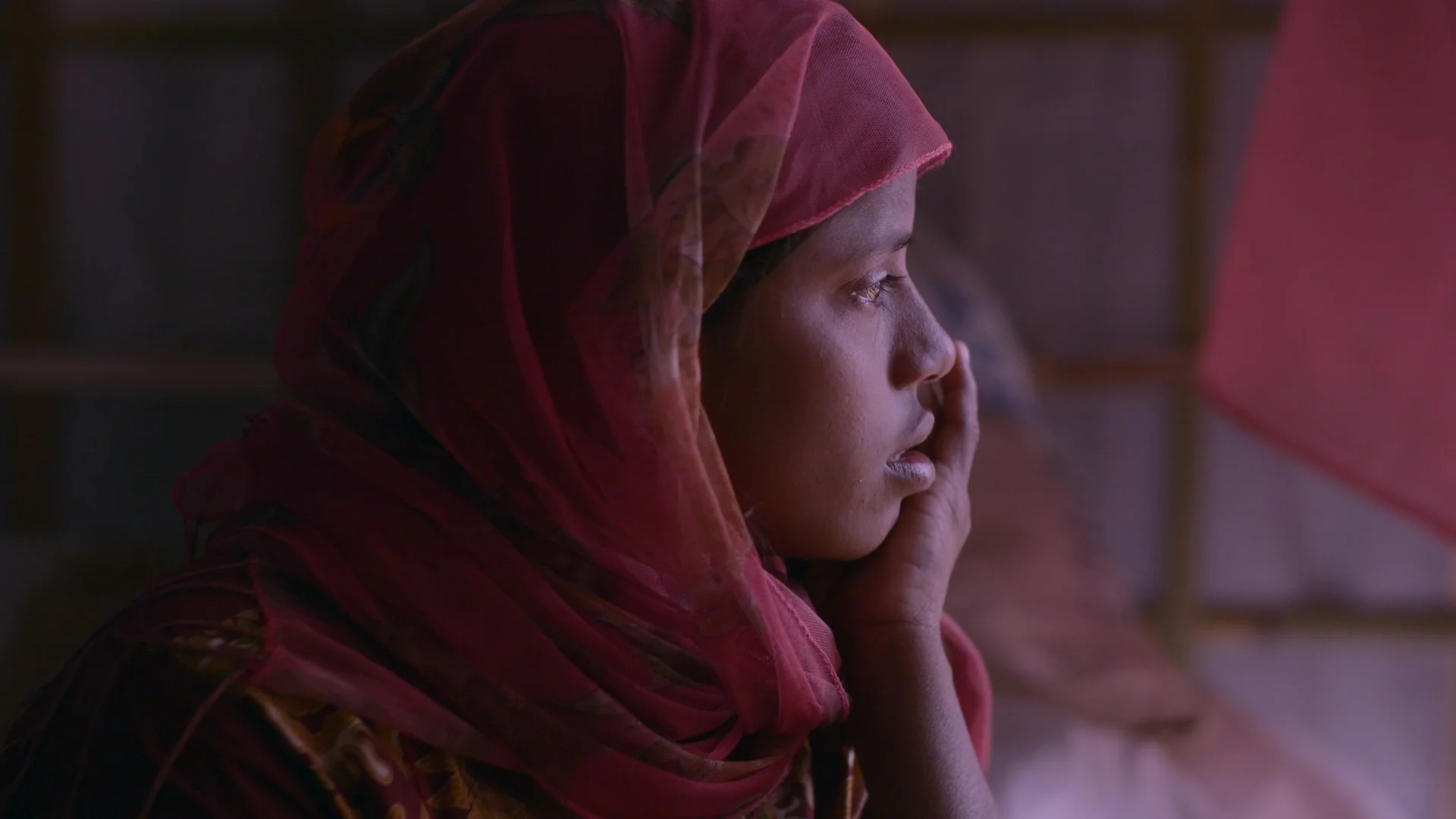 A woman with a dark complexion wearing a pink headscarf, looking contemplative with her hand resting on her face, in a dimly lit room.