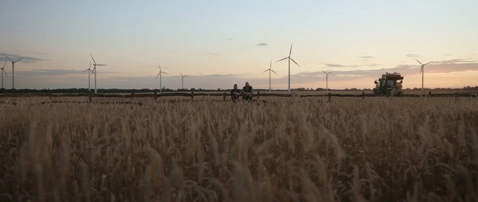 A rural farmland scene at sunset with a wheat field, wind turbines in the background, a tractor on the right, and two people sitting on a fence in the middle ground.