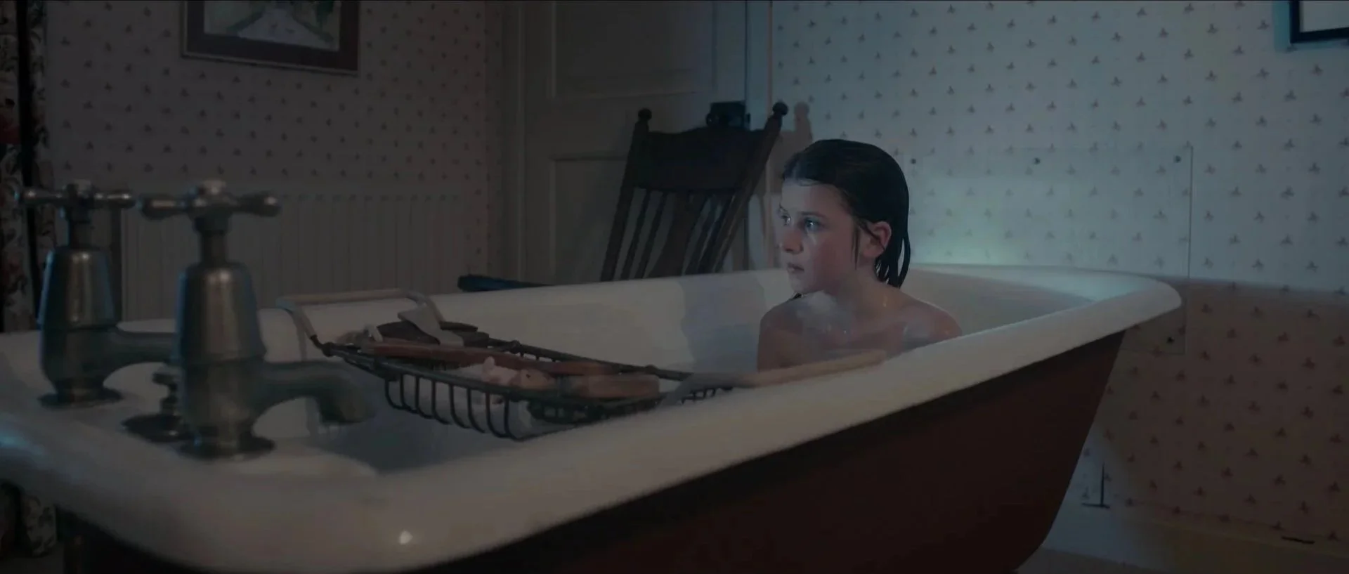 A young boy with dark hair sitting in a bathtub filled with water in a dimly lit bathroom, with a washing basket and fixtures visible in the foreground.