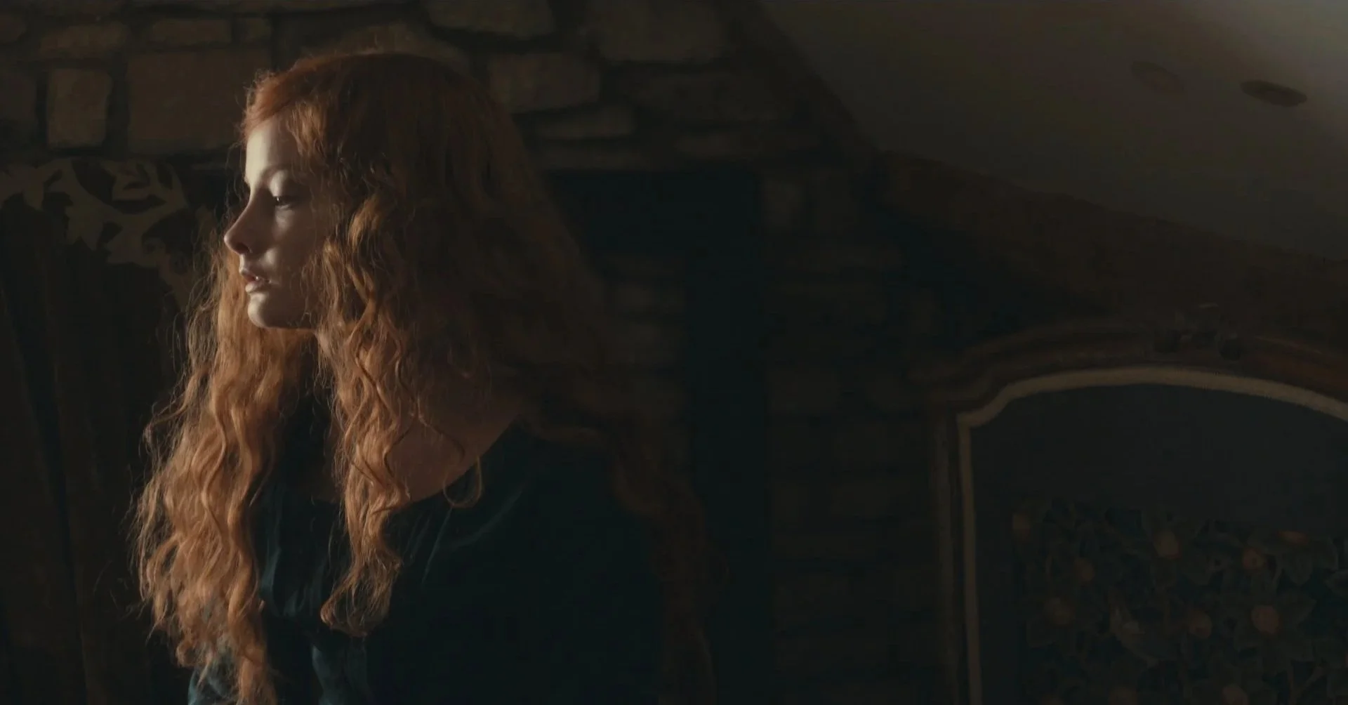 A woman with long, wavy red hair looking down in a dimly lit room with a stone wall in the background.