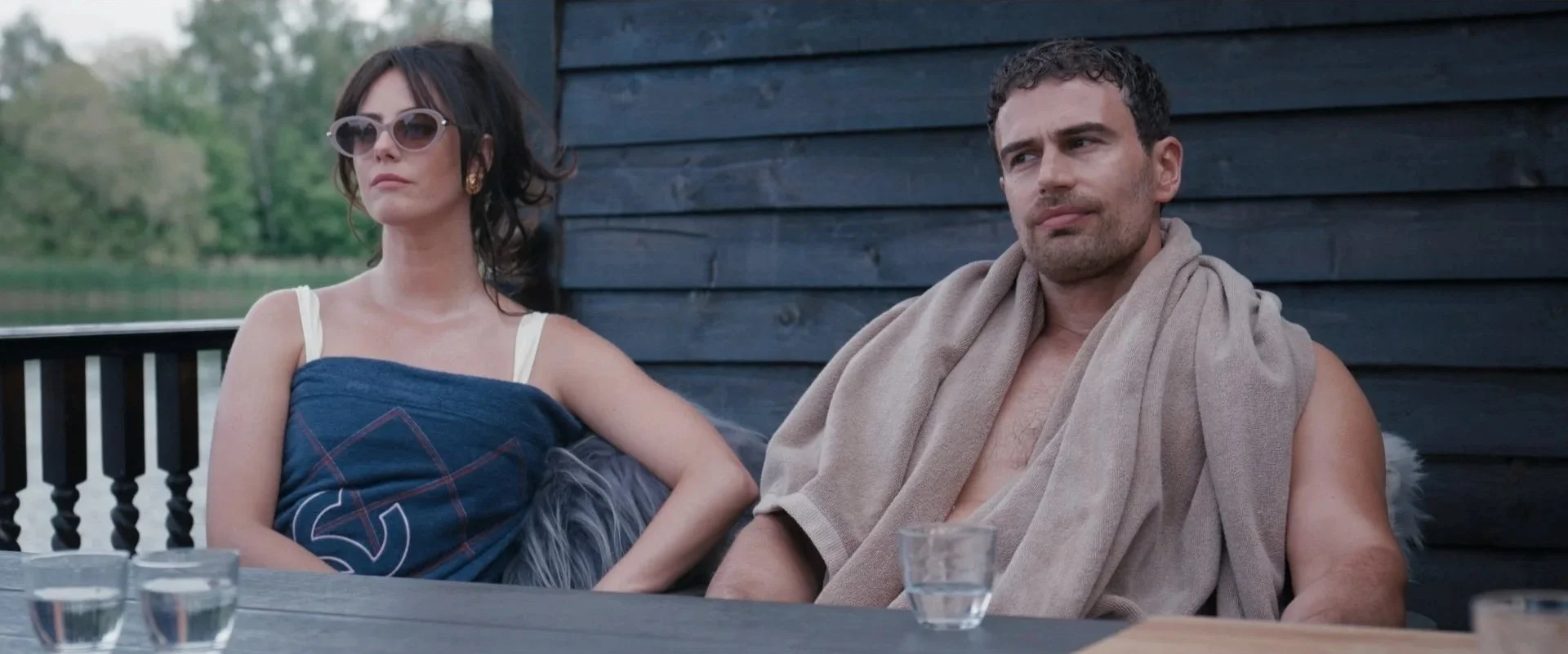 A woman wearing sunglasses and a sleeveless dress, and a man with a towel draped over his shoulders, sitting outdoors at a table with glasses of water, against a dark wooden wall with trees in the background.