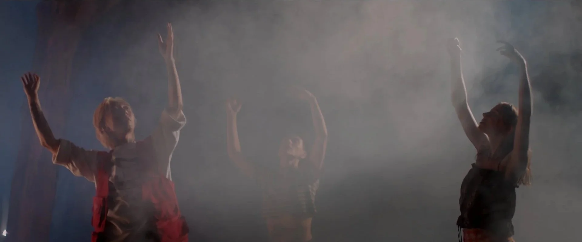 Three young children with arms raised, standing in a foggy environment with dramatic lighting, possibly performing or participating in an event or dance.