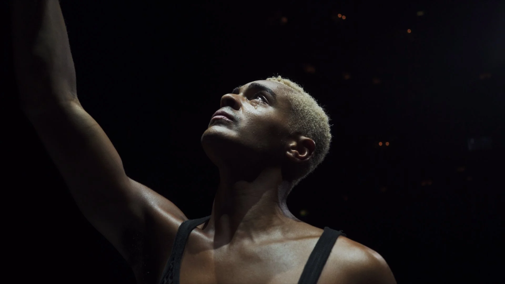 A person with short blonde hair looking upward with a tear on their cheek, in a dark setting with a black tank top.