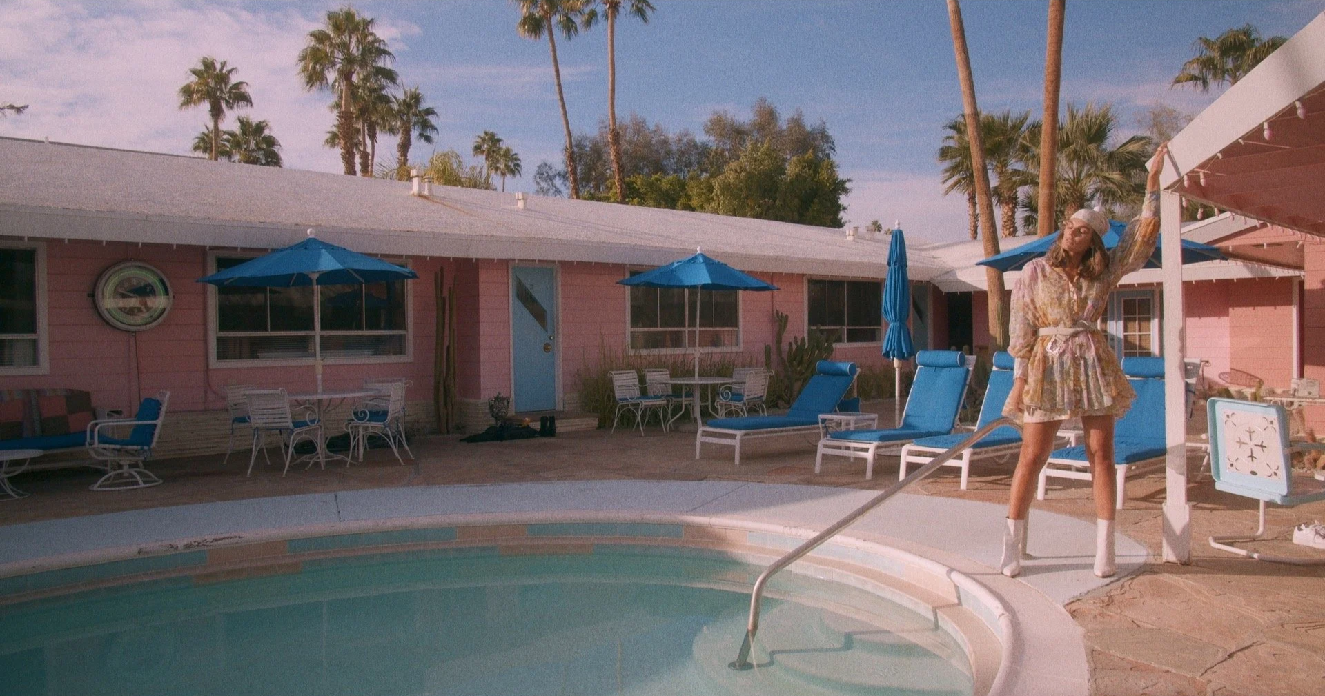 A woman in a floral dress and white boots standing by a swimming pool with pink house and blue patio furniture and umbrellas in the background, palm trees and a clear blue sky.