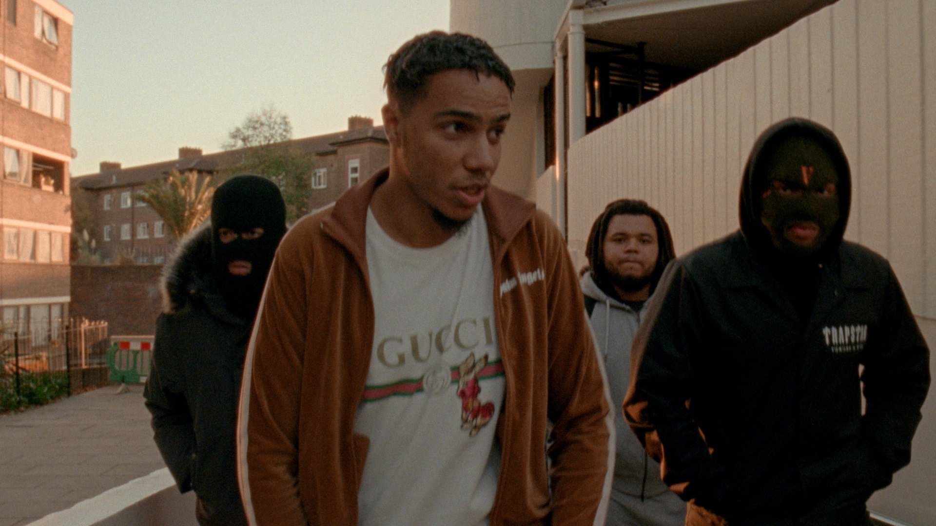 Four men walking outdoors in an urban area during sunset. Two men are wearing black ski masks and black jackets, and a young man in the center is wearing a brown jacket and a Gucci T-shirt. Behind them are residential buildings and a palm tree.