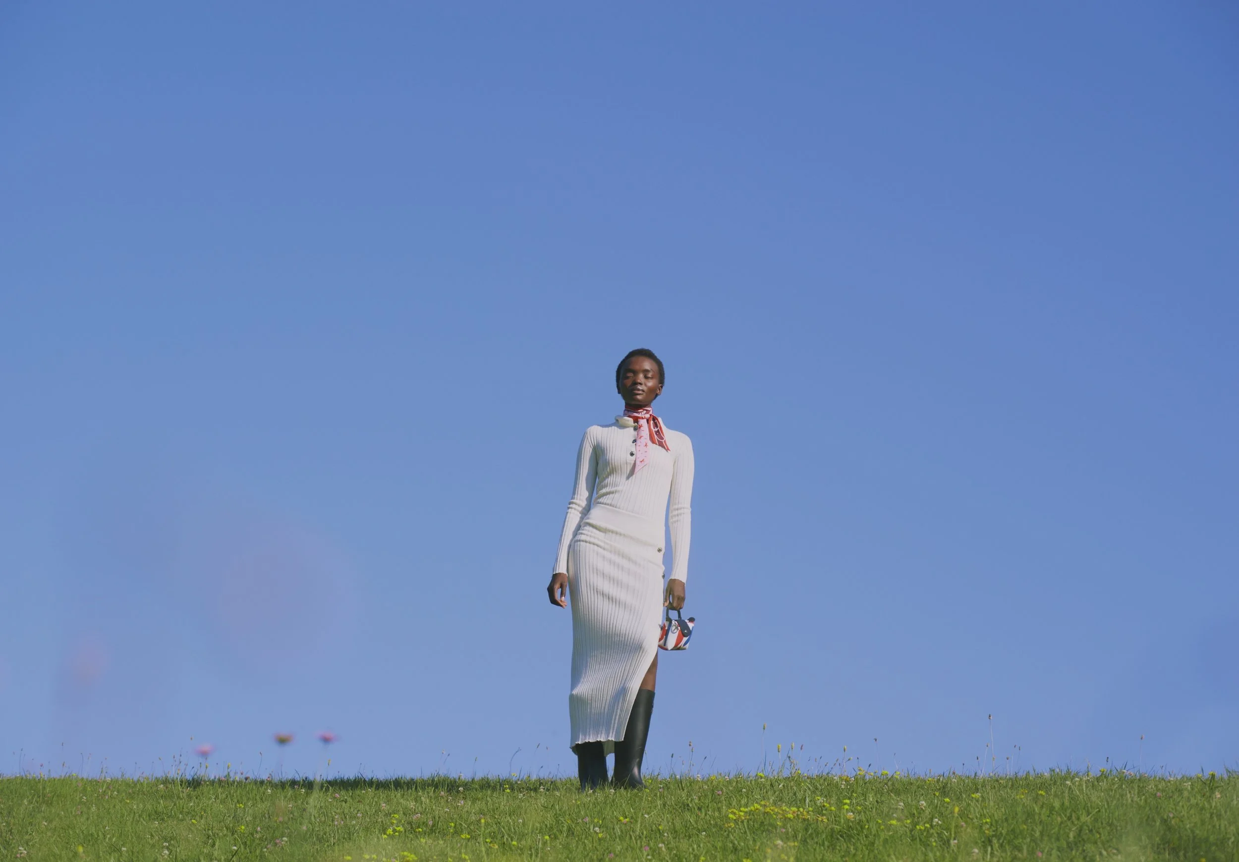 A woman standing in a grassy field under a clear blue sky, holding a small handbag, and wearing a white dress with black boots, with flowers visible in the background.