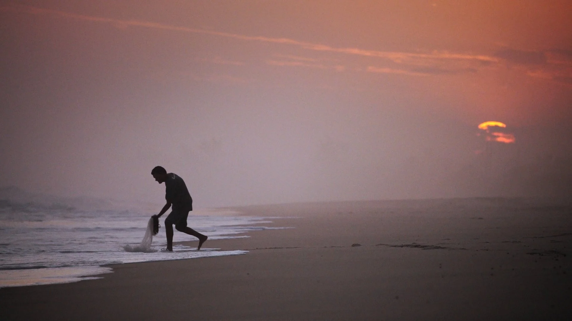 A person on the beach during sunset, bending down and holding a fishing net as the sun sets over the ocean.
