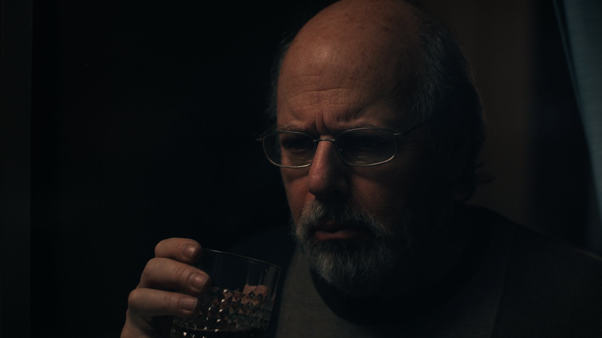 A middle-aged man with glasses and a beard looking at a glass of dark whiskey.
