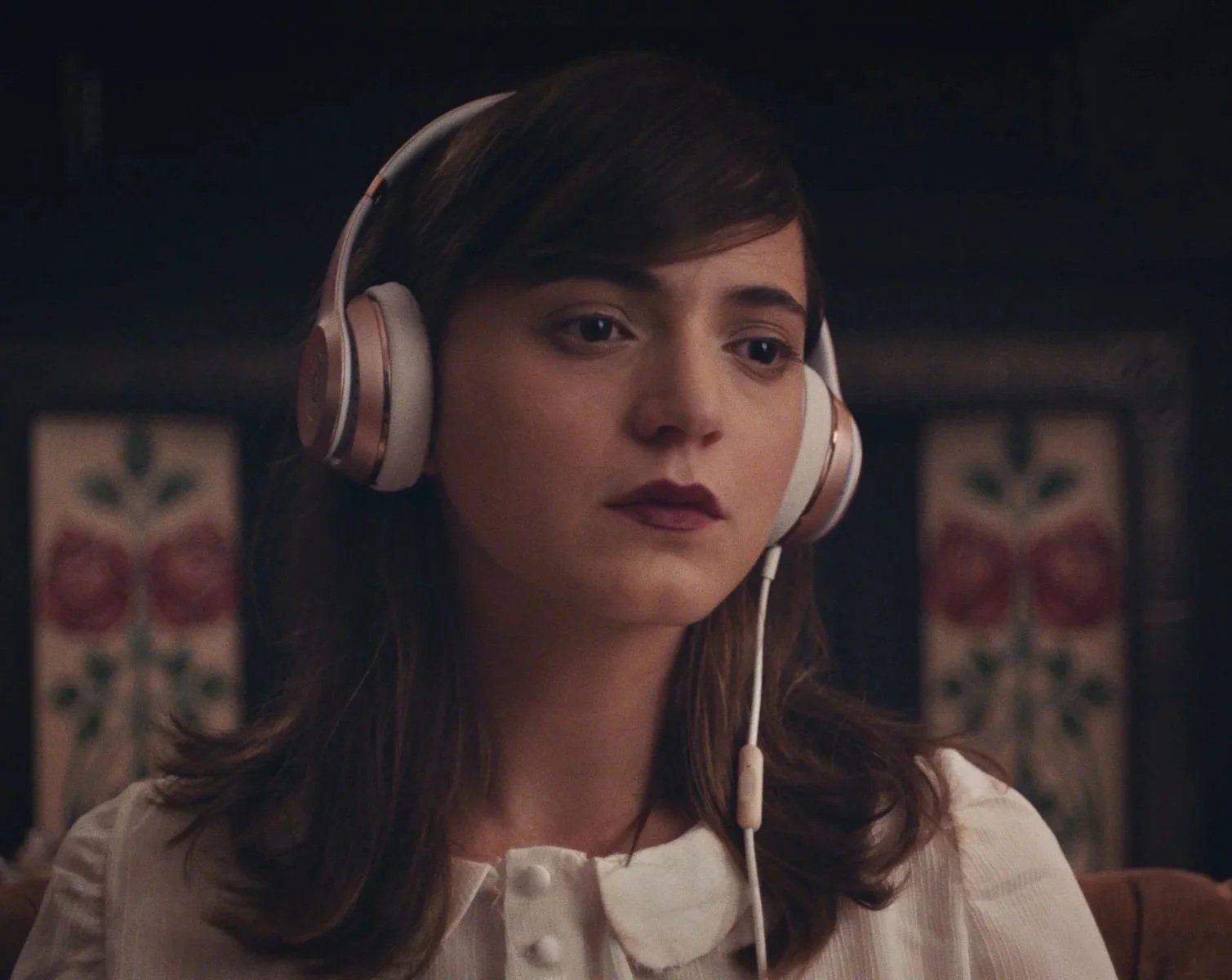 A woman with brown hair wearing headphones and a white blouse, looking thoughtful, indoors with blurred background featuring a patterned wall.